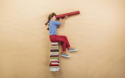 Girl sitting on a stack of books using rolled up cardboard as a telescope