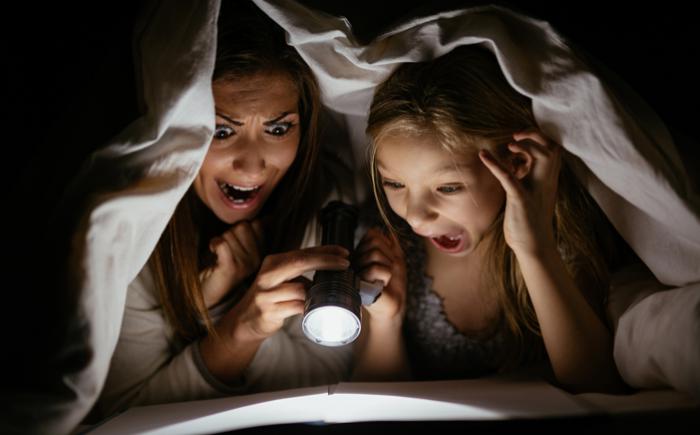 Mother and daughter reading a book by flashlight under a blanket