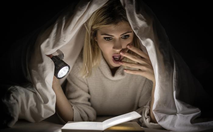 Woman reading scary book under cover with flashlight