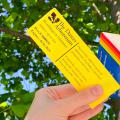 Hand holding Dawes Arboretum pass and library card against green leafy tree and blue sky