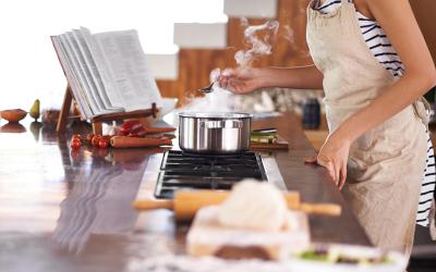 Woman wearing apron standing over stove with an open cookbook