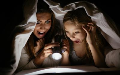 Mother and daughter reading a book under the covers with a flashlight
