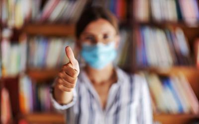 Woman in surgical mask in front of book shelves holding thumb up