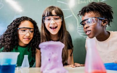 Three children wear safety glasses and watch liquid overflow a beaker