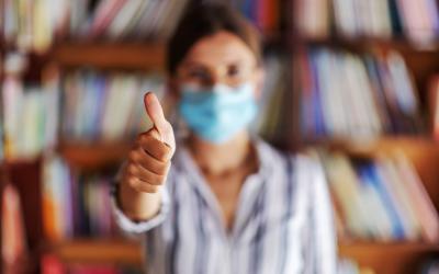 Woman standing in front of bookshelf, wearing a mask and giving a thumbs up
